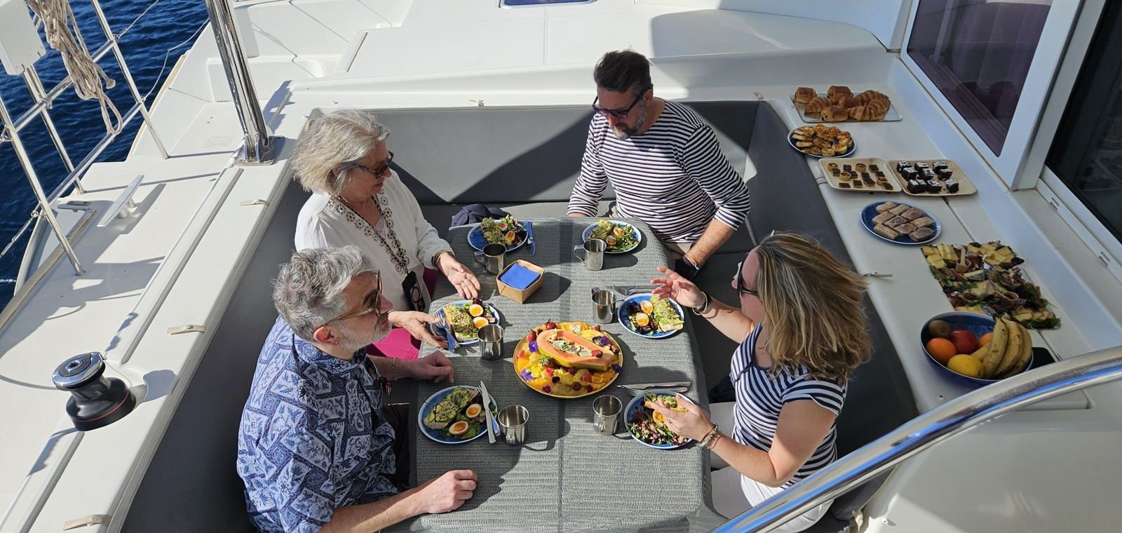 Quatre convives attablés sur le pont d'un catamaran, assiettes et planches dressées